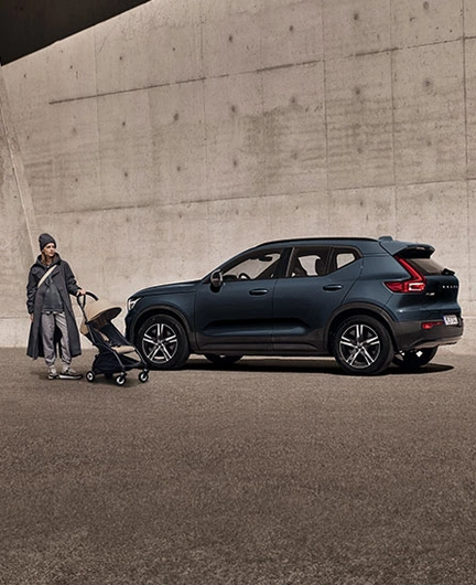 Une femme en imperméable tenant une poussette se tient debout devant un XC40 Volvo bleu