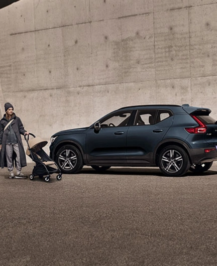 Une femme en imperméable tenant une poussette se tient debout devant un XC40 Volvo bleu