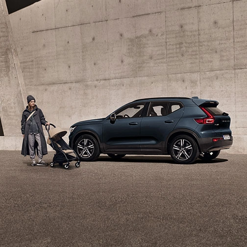 Une femme en imperméable tenant une poussette se tient debout devant un XC40 Volvo bleu