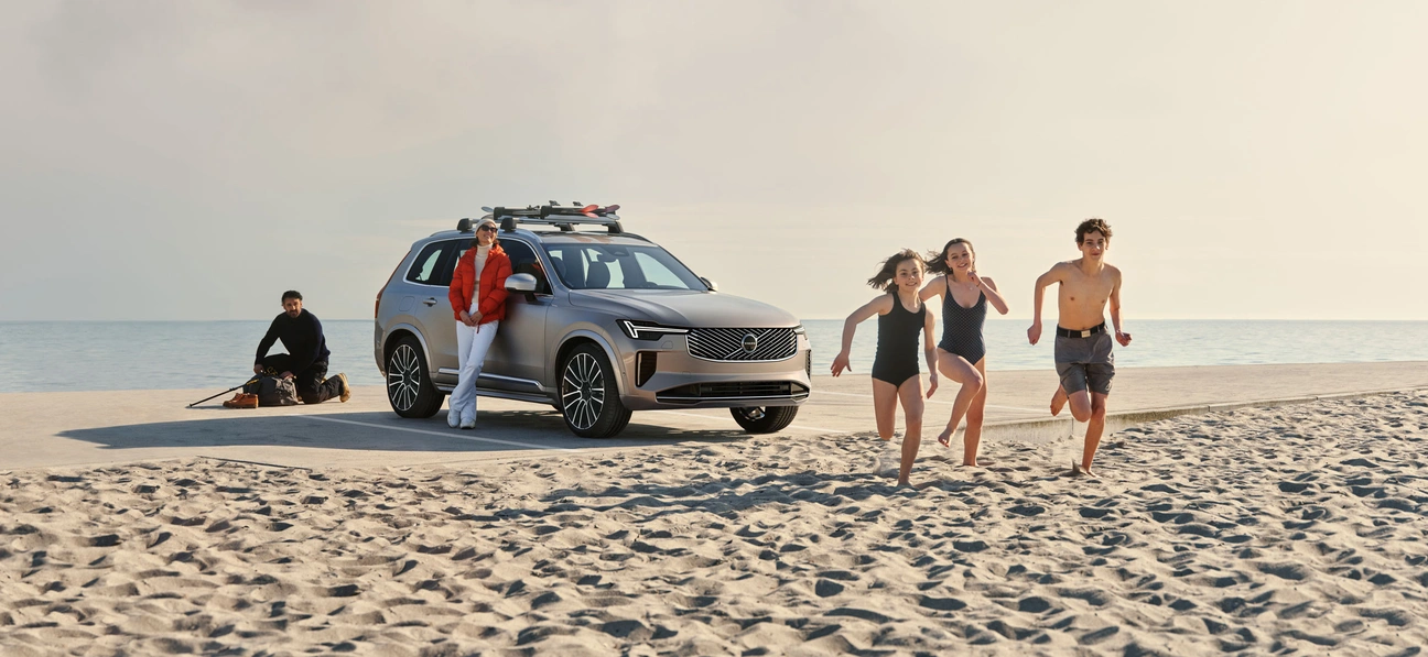Voiture Volvo XC90 sur une plage, personne appuyée contre elle, trois enfants courant sur le sable.