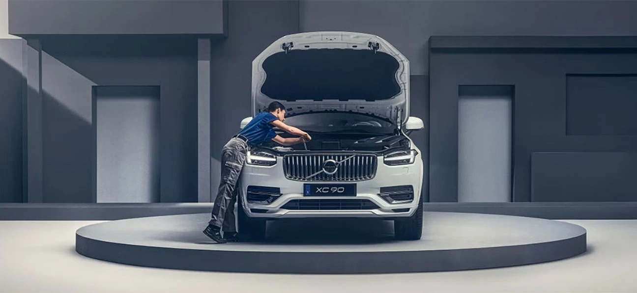 Personne inspectant le moteur d’un Volvo XC90 blanc sur une plateforme circulaire dans un décor minimaliste