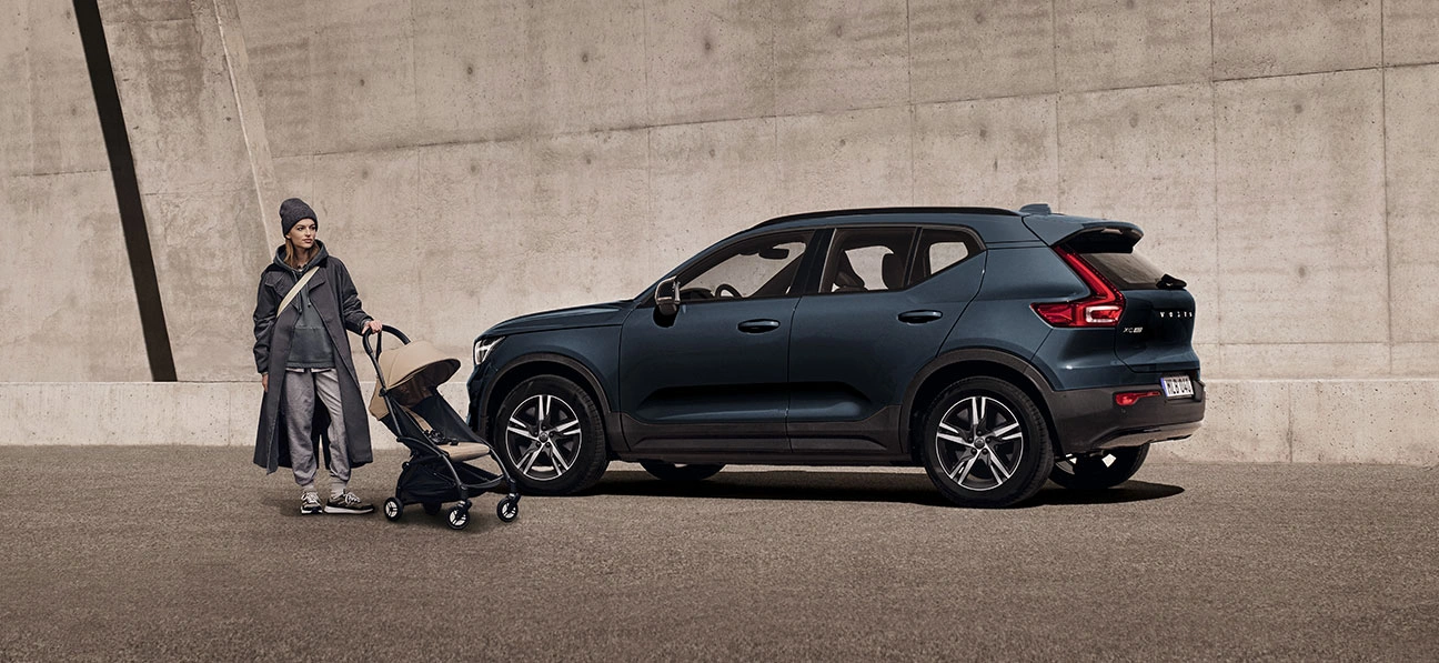 Une femme en imperméable tenant une poussette se tient debout devant un XC40 Volvo bleu