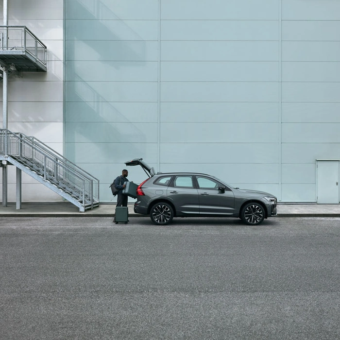 Homme chargeant une valise dans le coffre d’un SUV XC60 gris, garé devant un bâtiment avec escalier métallique.