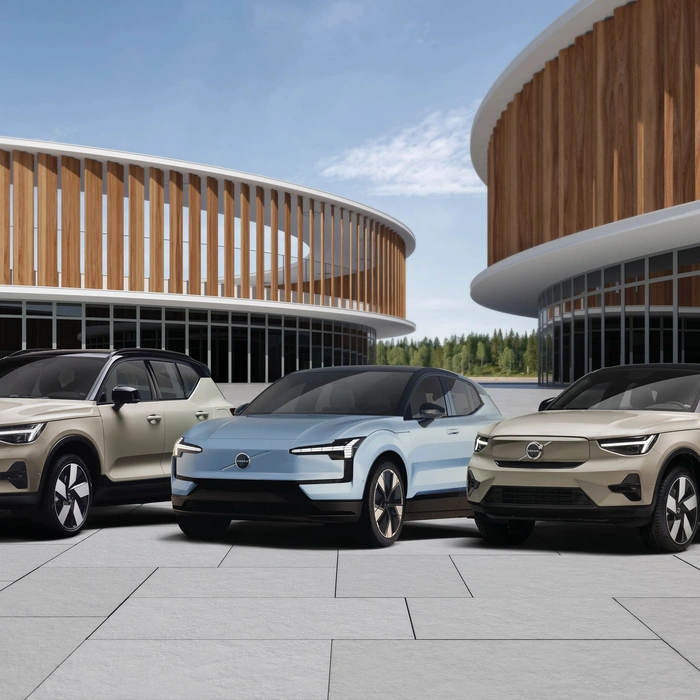Trois voitures électriques Volvo garées côte à côte sur une grande esplanade en béton. À gauche et à droite, deux SUV couleur beige, et au centre une berline bleu clair. En arrière-plan, deux bâtiments circulaires modernes avec des façades en bois et de grandes baies vitrées.